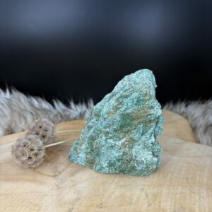 Fuchsiet natuursteen van 11 cm hoog met een zachte groene glans, Detail van de glinstering in een Fuchsiet van 334 gram, Zijaanzicht van de 3 cm diepe Fuchsiet met zilverachtige mica schittering, Groene Fuchsiet op een houten ondergrond in een rustige woonkamer.