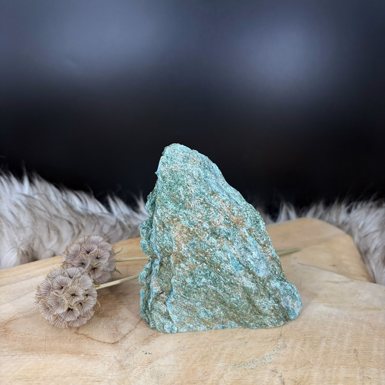 Fuchsiet natuursteen van 11 cm hoog met een zachte groene glans, Detail van de glinstering in een Fuchsiet van 334 gram, Zijaanzicht van de 3 cm diepe Fuchsiet met zilverachtige mica schittering, Groene Fuchsiet op een houten ondergrond in een rustige woonkamer.