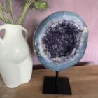 Amethist Geode op standaard, Een glanzende Amethist Geode op zwarte standaard, close-up van de kristallen in de Amethist Geode, zijkant van een grote Amethist Geode met diepe paarse punten, sfeervolle woonkamer met Amethist Geode als decoratie, detailfoto van glinsterende amethistkristallen, Amethist Geode op standaard bij een meditatiehoek, natuurlijk gevormde Amethist Geode met unieke structuur, Amethist Geode op standaard met zachte verlichting eromheen, overzicht van verschillende Amethist Geodes op standaard in een interieursetting.