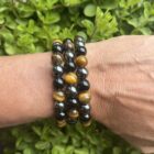 Armband van hematiet, obsidiaan en tijgeroog op houten altaar, Detail van 8 mm kralen in aardende kleurcombinatie, Pols met krachtige drievoudige edelsteenarmband, Spirituele setting met hematiet-obsidiaan-tijgeroog armband, Armband naast palo santo en seleniet bij volle maanritueel