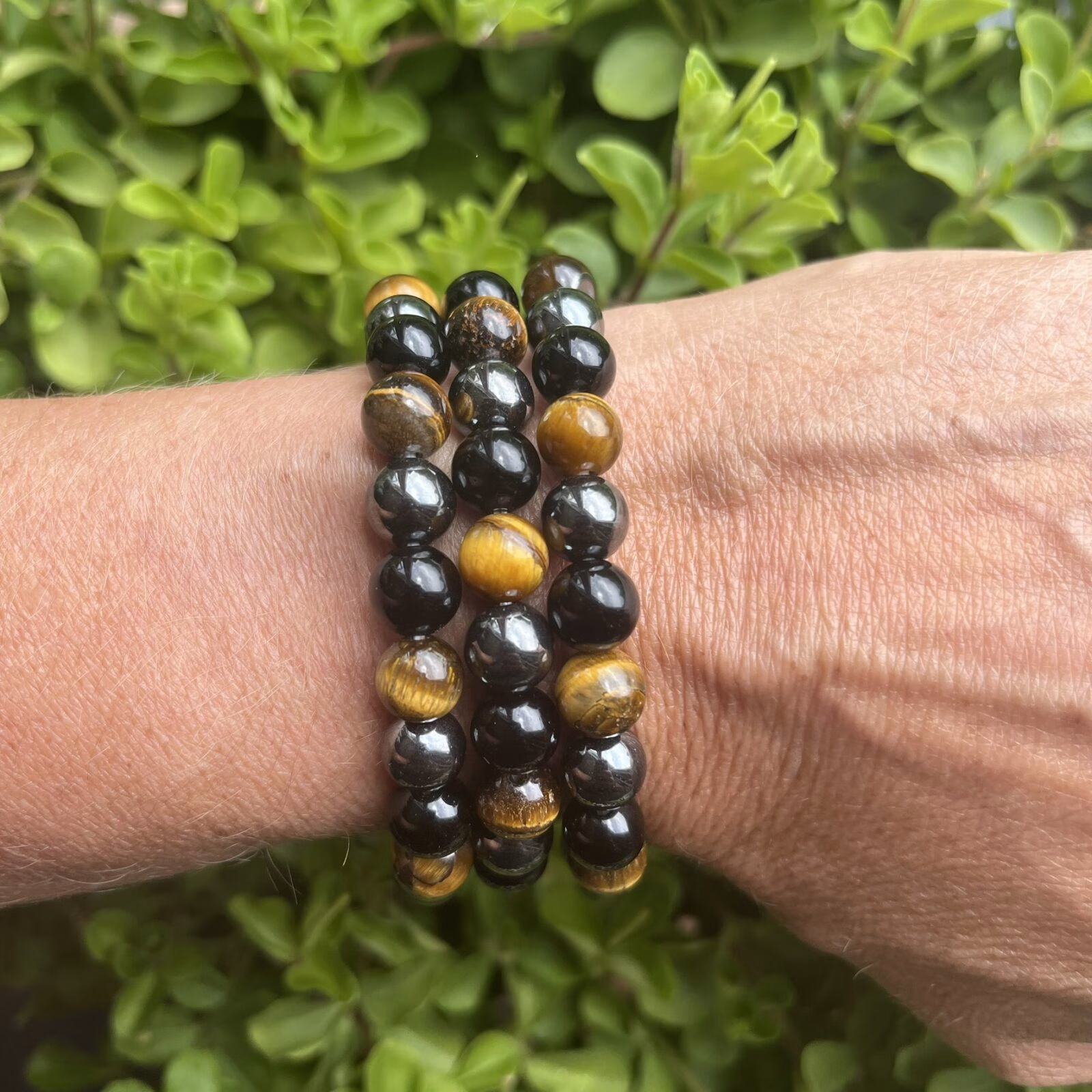 Armband van hematiet, obsidiaan en tijgeroog op houten altaar, Detail van 8 mm kralen in aardende kleurcombinatie, Pols met krachtige drievoudige edelsteenarmband, Spirituele setting met hematiet-obsidiaan-tijgeroog armband, Armband naast palo santo en seleniet bij volle maanritueel