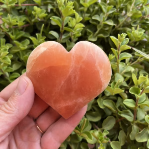 oranje seleniet hart met warme gloed op houten tafel, close-up van oranje kristal in hartvorm met zachte randen, spiritueel altaar met seleniet hart en kaarslicht, 7 cm oranje seleniet op vensterbank in zonsondergang, hand houdt hartvormige oranje steen tegen rustige achtergrond
