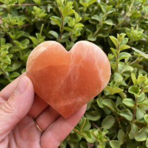 oranje seleniet hart met warme gloed op houten tafel, close-up van oranje kristal in hartvorm met zachte randen, spiritueel altaar met seleniet hart en kaarslicht, 7 cm oranje seleniet op vensterbank in zonsondergang, hand houdt hartvormige oranje steen tegen rustige achtergrond