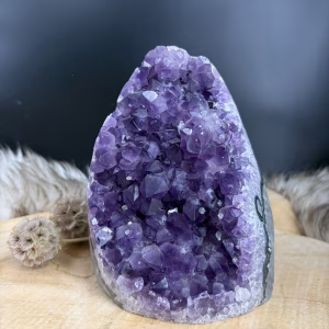 Vooraanzicht van een dieppaarse amethist cut-base uit Uruguay van 2118 gram. Detailopname van de glanzende paarse kristalpunten van de ruwe amethist. Zijaanzicht van de amethist met de natuurlijke grijze achterkant en rechte onderzijde. De amethist uit Uruguay geplaatst op een houten tafel in een lichte woonkamer.