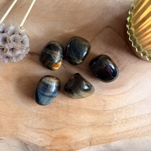 Valkenoog Trommelsteen, Valkenoog Edelsteen, Valkenoog trommelsteen op houten plank met kaarsje, Valkenoog trommelsteen 20 gram voor rust en overzicht, blauwe Tijgeroog edelsteen met zijdeglans bij Natuursteentje, handgeselecteerde Valkenoog kopen voor focus, spirituele werking Valkenoog tegen stress, edelsteen combinatie met Tijgeroog en Lapis Lazuli, unieke blauwgrijze trommelsteen webshop Natuursteentje.