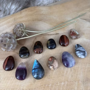 Een groep doorboorde edelsteen hangers van Labradoriet, Mahonie Obsidiaan, Pinoliet, Regenboog Fluoriet, Rode Tijgeroog, Valkenoog en Poppy Jaspis op een houten ondergrond
